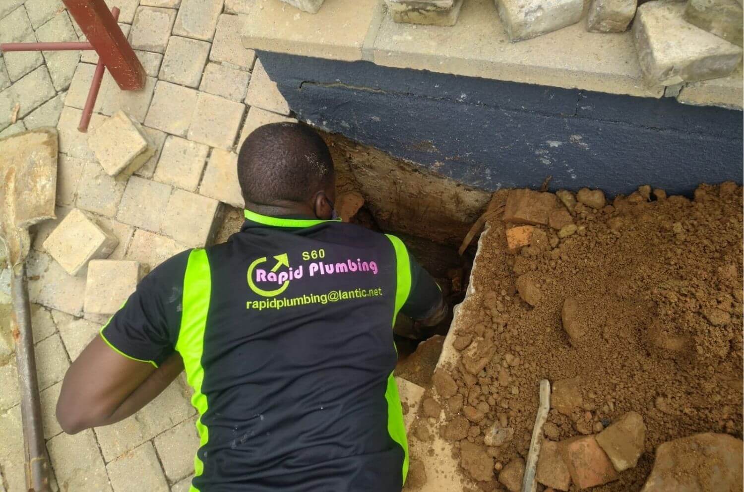 A professional technician wearing a Rapid Plumbing uniform while performing a manual drain excavation and repair in a residential backyard